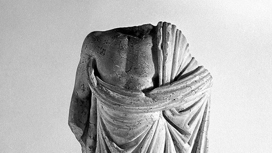 Black-and-white photo an Ancient Greek statue of a man in a toga, cropped to show the figure from roughly the waist to above the shoulders. It is headless, the neck broken off the body