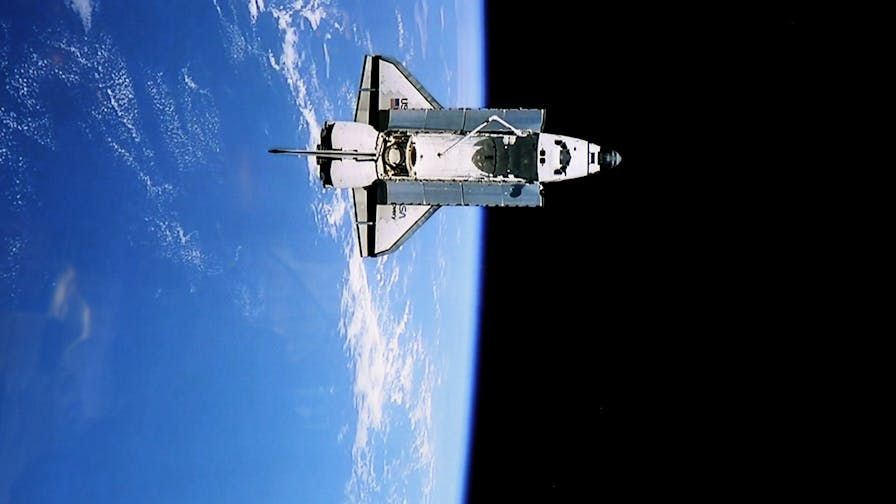 Color photo of a space shuttle in space, showing the top of the shuttle and, in the background, Earth