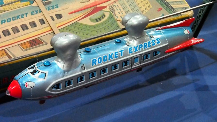 Color photo of an antique toy commuter rocketship with windows down the side like an airplan. It is painted metalic silver and blue with red fins and nose cone, and is labelled Rocket Express. In the background is its box.